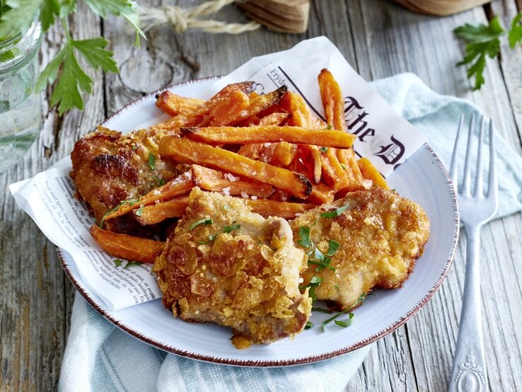 Crisp-Schnitzel mit Süßkartoffelpommes auf einem Teller. Rechts eine Gabel, links davon Petersilie. Draufsicht.