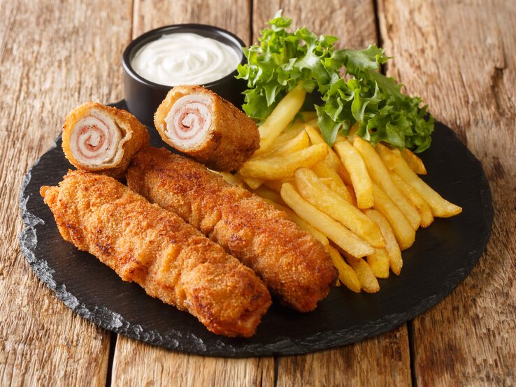 Flamenquín mit Pommes, Salat und Mayonnaise auf Schieferplatte, positioniert auf hölzernem Untergrund. 