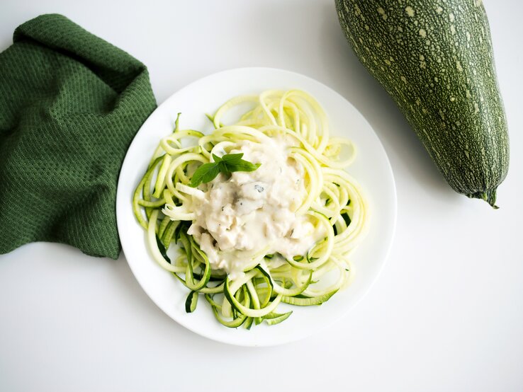 Auf einem weißen Untergrund liegen ein grünes Tuch und eine Zucchini. Daneben steht ein weißer Teller mit Zucchini-Nudeln mit Ziegenkäse-Soße.