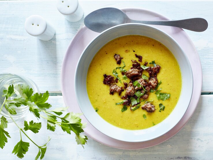 Auf einem Untergrund aus Holz stehen ein Salz und ein Pfefferstreuer. Daneben ein Glas mit einem Zweig Petersilie. Und auch ein Teller auf dem eine Suppenschüssel mit einer Currysuppe mit Hackfleisch   steht. Neben der Schüssel liegt ein Löffel.