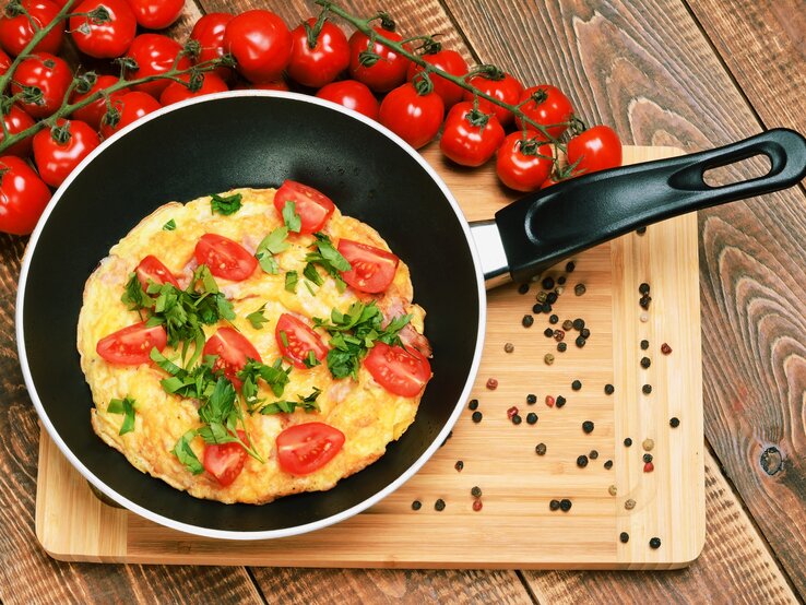 Eine Pfanne mit dem Tomatenomelett auf einem Holztisch mit einem Strauch Kirschtomaten von oben fotografiert.