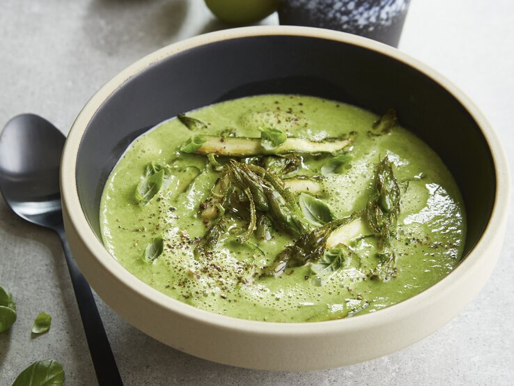 Diese leichte Spargelsuppe aus grünem Spargel und Limette passt perfekt in den Frühling! Eine graue Schale auf einem grauen Tisch mit der grünen Spargelcemesuppe. Alles von schräg oben fotografiert.