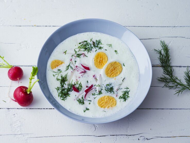 Kalte Sommersuppe mit Joghurt, Radieschen, Dill und hartgekochten Eiern in einer blauen Schale auf weißem Holztisch. Alles direkt von oben fotografiert.