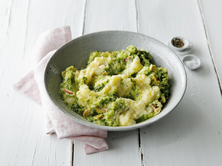 Eine weiße Schüssel mit dem Kartoffelstampf auf einem weißen Holztisch. Alles von schräg oben fotografiert.