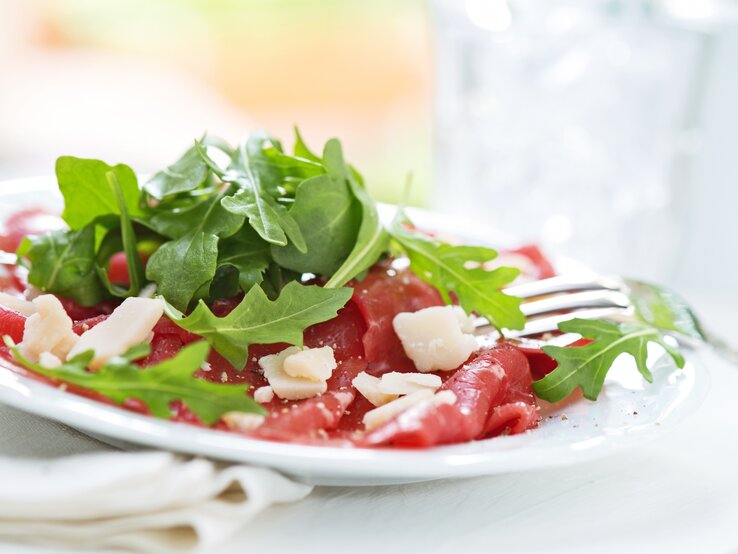 Rinder-Carpaccio mit Parmesan und Rucola. Köstlich, leicht, original italienisch! Seitenansicht auf einen weißen Teller belegt mit Rinder-Carpaccio