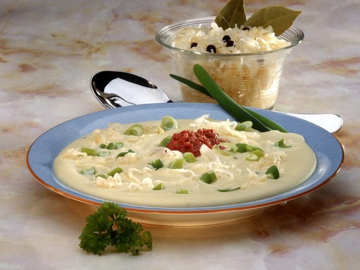 Eine Draufsicht auf einen Teller mit cremiger Suppe, garniert mit frischen grünen Zwiebelringen, geriebenem Käse und einem Klecks roter Soße in der Mitte. Die Suppe wird in einem blauen Suppenteller mit einem orangefarbenen Rand serviert. Im Hintergrund befindet sich eine Glasschale mit weiterem geriebenem Käse, verziert mit Lorbeerblättern und Pfefferkörnern. Die Szene liegt auf einer Marmor-Tischoberfläche und wird von einem silbernen Löffel und einem frischen Petersilienzweig ergänzt. 