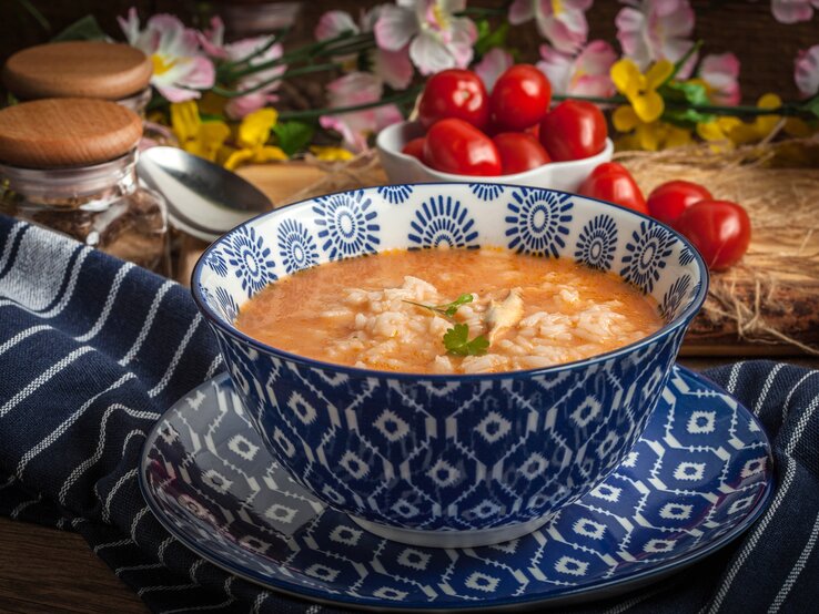 Eine Schüssel Tomatensuppe mit Reis, serviert in einer tiefblauen, mit weißen Mustern verzierten Schale auf einem Untersetzer gleichen Designs. Die Schale steht auf einem dunkelblauen Tuch mit weißen Streifen. Im Hintergrund sind Gewürzgläser, ein Löffel und frische Kirschtomaten zu sehen, umgeben von rustikalen Holzelementen und Frühlingsblumen.