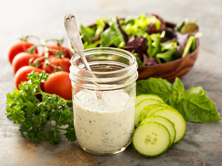 Frisch angerührtes Joghurtdressing mit frischen Kräutern im Glas. Daneben liegen knackige Salatzutaten.