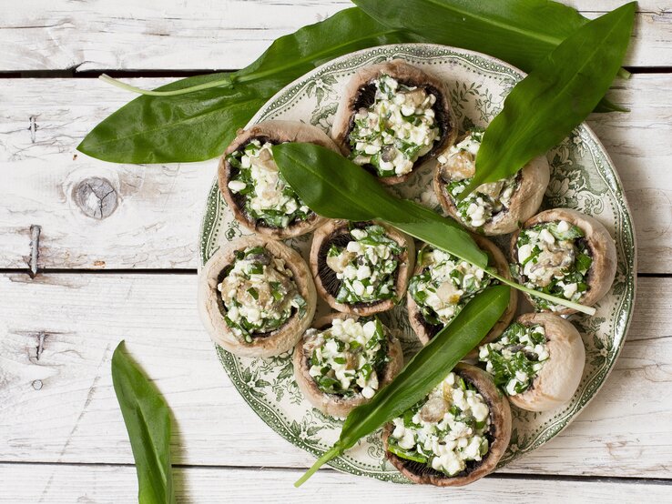 Gefüllte Champignons mit einer Füllung aus Bärlauch und Feta auf einem hellen verzierten Teller mit ganzen Bärlauch-Blättern auf einem hellen Holztisch von oben.