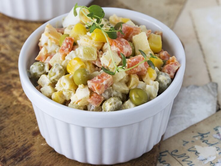 Eine kleine weiße Schale gefüllt mit einem Kartoffelsalat mit Mais, Ei, Erbsen, Karotten und Mayonnaise.