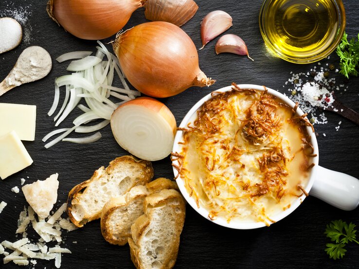  Das Bild zeigt eine appetitlich angerichtete französische Zwiebelsuppe in einem weißen Keramiktopf. Die Suppe ist mit geschmolzenem Käse überbacken, der goldbraun gratiniert ist. Um den Topf herum sind die Zutaten für die Suppe verteilt: frische Zwiebeln, geschnittene Zwiebelringe, Knoblauchzehen, ein Stück Butter, Olivenöl, eine Prise Salz und Pfeffer, sowie frisches Brot und geriebener Käse.