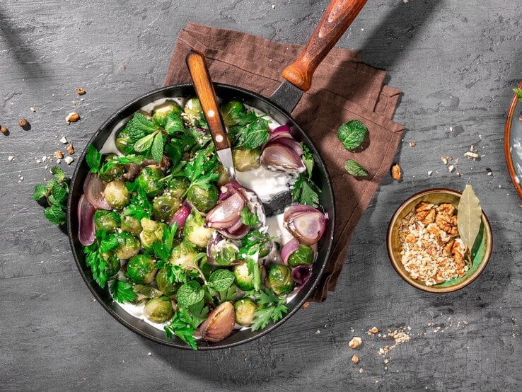 Draufsicht auf eine runde Pfanne mit hölzernem Stiel. In der pfanne ist gebratener Rosenkohl und Joghurtsoße. 