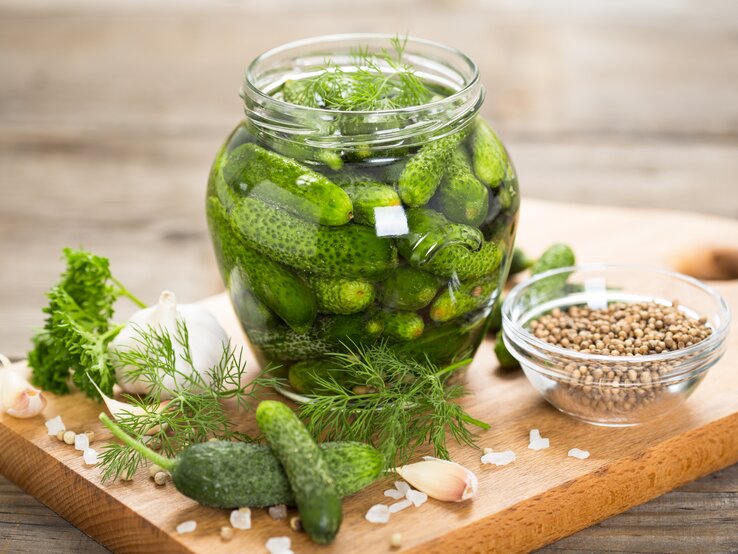 Einmachglas gefüllt mit Gewürzgurken. Daneben liegen frischer Dill, Senfkörner, Knoblauchzehen und Meersalz.