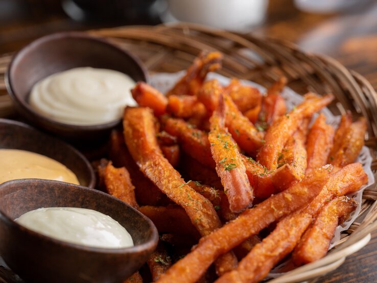 Suesskartoffelpommes-Heissluftfritteuse Süßkartoffelpommes aus der Heißluftfritteuse in Bambusschale mit dreierlei Dips in braunen Schalen auf Holztisch. Draufsicht.