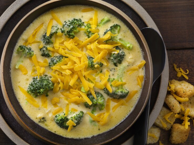 Das Bild zeigt eine Schale mit Brokkoli-Käse-Suppe. Die Suppe hat eine cremige Konsistenz und ist mit viel geriebenem Cheddar-Käse bestreut, was für eine reiche und ansprechende Textur sorgt. Die leuchtend grünen Brokkoliröschen schwimmen in der gelblichen Suppe und bieten einen schönen Farbkontrast. Am Rand der Schale sind einige Croutons zu sehen, die als Beilage dienen könnten. Der dunkle Holzhintergrund verleiht dem Bild Wärme und betont die appetitliche Präsentation der Speise.