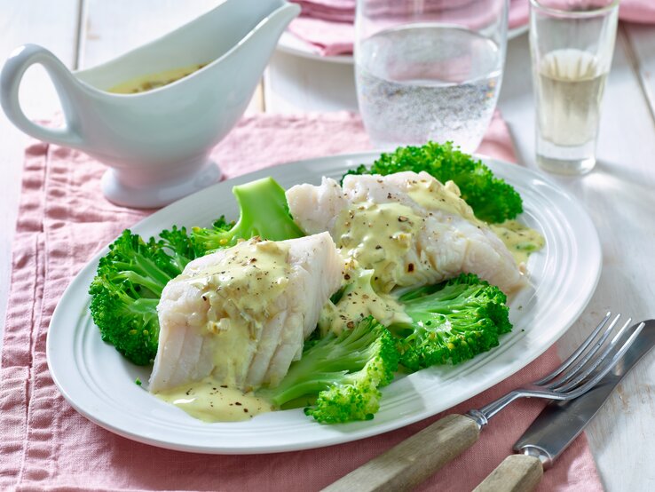 Zanderfilet mit Brokkoli auf einem runden Teller aus Porzellan / Messer und Gabel / ein Behältnis aus Porzellan mit Sauce / Ein Glas mit Sprudelwasser 