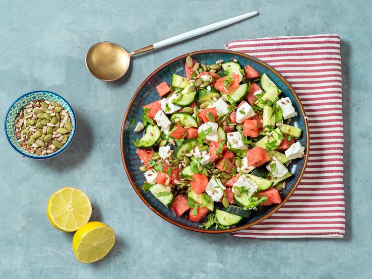 Gurkensalat mit Wassermelone und Feta auf blauem Teller, rot-weißem Tuch und hellblauem Untergrund. Im BIld eine Lössel, eine Schale mit Kernen und eine geteilte Zitrone. Draufsicht.