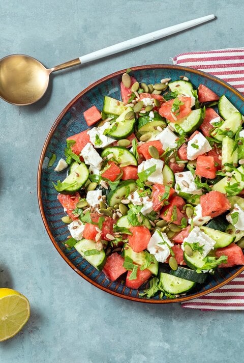 Gurkensalat mit Wassermelone und Feta auf blauem Teller, rot-weißem Tuch und hellblauem Untergrund. Im BIld eine Lössel, eine Schale mit Kernen und eine geteilte Zitrone. Draufsicht.