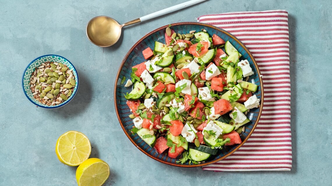 Gurkensalat mit Wassermelone und Feta auf blauem Teller, rot-weißem Tuch und hellblauem Untergrund. Im BIld eine Lössel, eine Schale mit Kernen und eine geteilte Zitrone. Draufsicht.