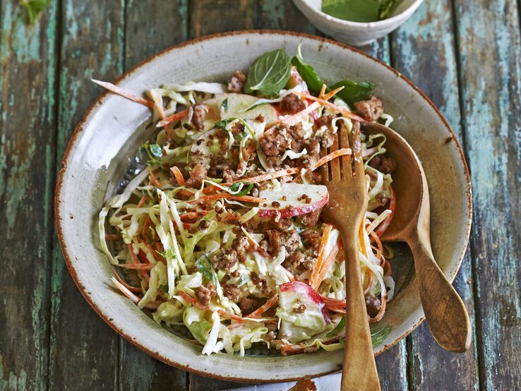 Möhren-Spitzkohl-Salat mit Rinderhack und Apfel / runder Teller / Löffel und Gabel aus Holz / Untergrund Holzplanken / Draufsicht, schräg