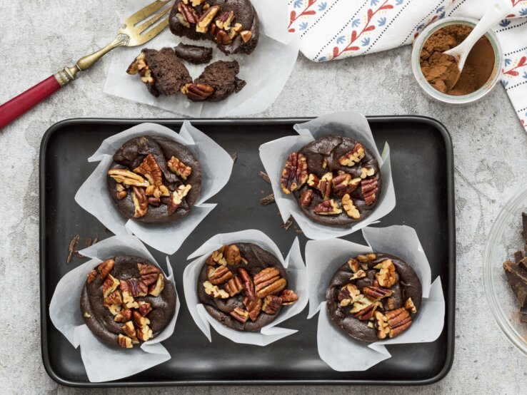 Schoko-Muffins-Avocado Die Schokomuffins mit Avocado und Nüssen auf einem Blech und grauem Tisch. Alles direkt von oben fotografiert.