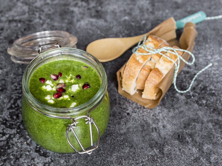 Grünkohlsuppe mit Granatapfel in einem Bügelverschlussglas vor rustikalem Hintergrund. Rechts Baguettte-Scheiben. Draufsicht.