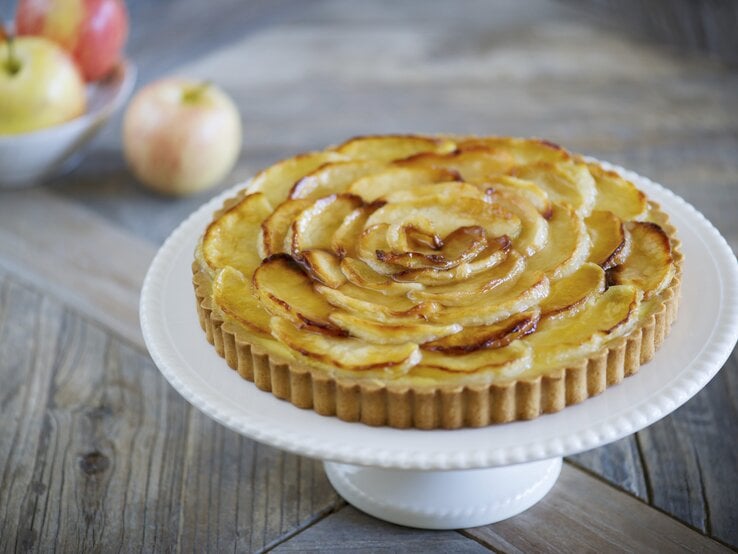 Die Apfel-Tarte auf einem weißen Tortenständer und rustikalem Holztisch von schräg oben fotografiert.