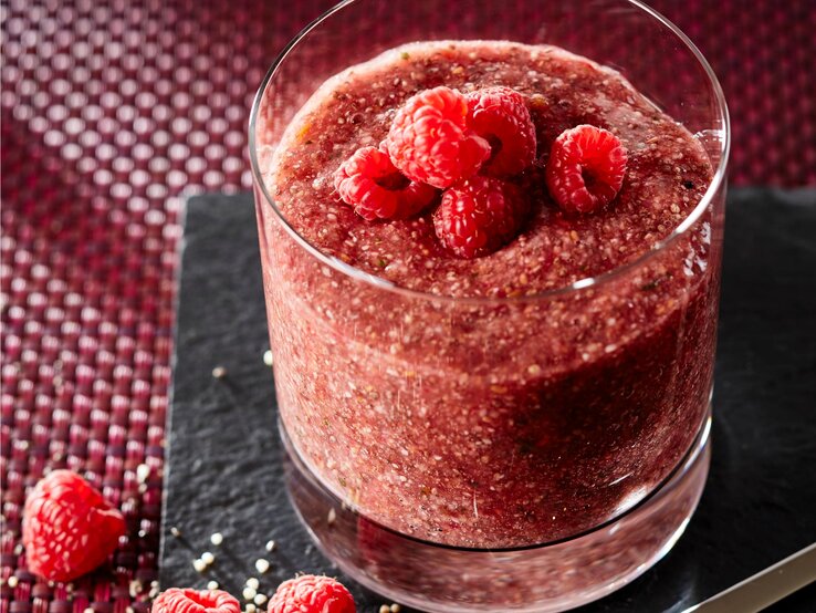 Das Bild zeigt ein Glas gefüllt Himbeer-Chia-Pudding. Auf der Oberfläche des Getränks befinden sich drei ganze Himbeeren als Garnierung. 