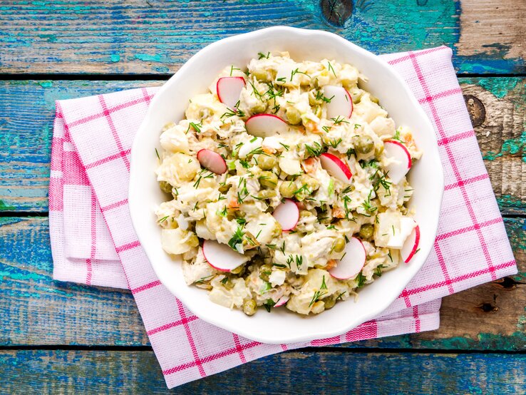 Falscher Kartoffelsalat mit Radieschen und Kräutern in weißer Schale auf rot-weißer Serviette, rustikaler Holztisch.