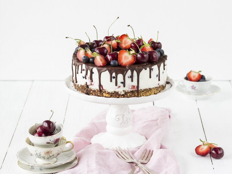 Weiße Beerentorte mit Kirschen und Erdbeeren auf einem Tortenständer, mit Schokoladenglasur am Rand. Die Torte steht auf einem hellen Holztisch, umgeben von Vintage-Tassen, Besteck und einem rosa Tuch.