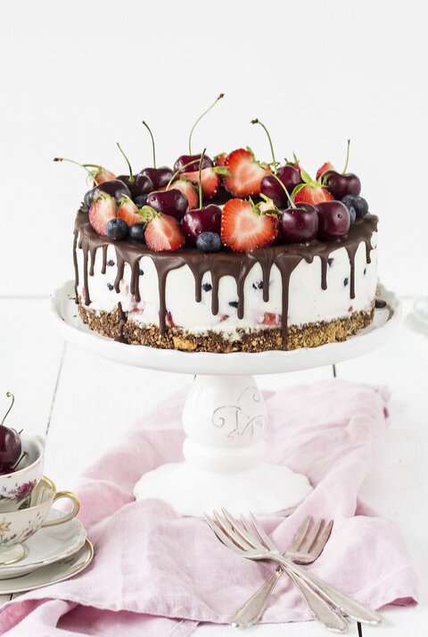Weiße Beerentorte mit Kirschen und Erdbeeren auf einem Tortenständer, mit Schokoladenglasur am Rand. Die Torte steht auf einem hellen Holztisch, umgeben von Vintage-Tassen, Besteck und einem rosa Tuch.