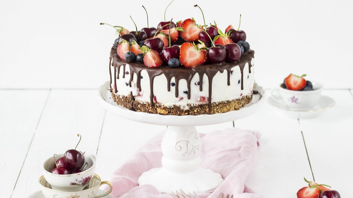 Weiße Beerentorte mit Kirschen und Erdbeeren auf einem Tortenständer, mit Schokoladenglasur am Rand. Die Torte steht auf einem hellen Holztisch, umgeben von Vintage-Tassen, Besteck und einem rosa Tuch.