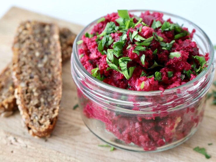 Nahaufnahme vom Rote-Bete-Aufstrich in einem Schraubglas, daneben zwei dunkle Brotscheiben.