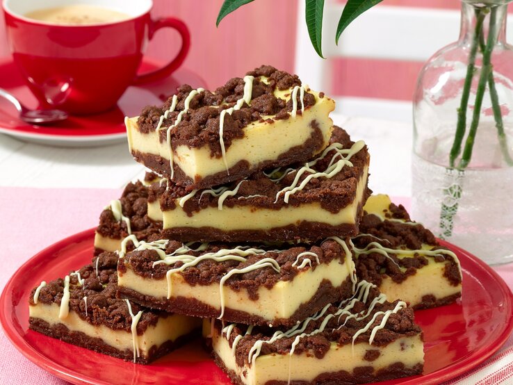 Mehrere quadratische Stücke Käsekuchen mit dunklen Schokostreuseln sind auf einem roten Teller gestapelt.