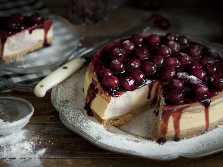 Kaesekuchen-Kirschen Der angeschnittene Käsekuchen auf einem dunklen Teller in rustikalem Ambiente.