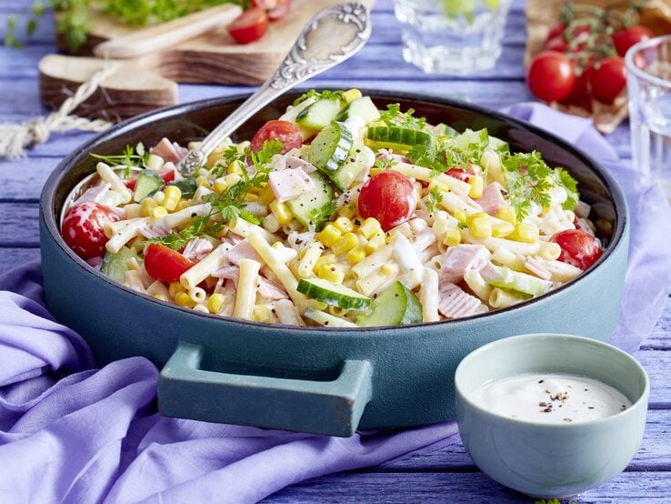 Nudelsalat mit Schinken in einer blauen Schüssel. Daneben ein Dressing und andere Zutaten. 