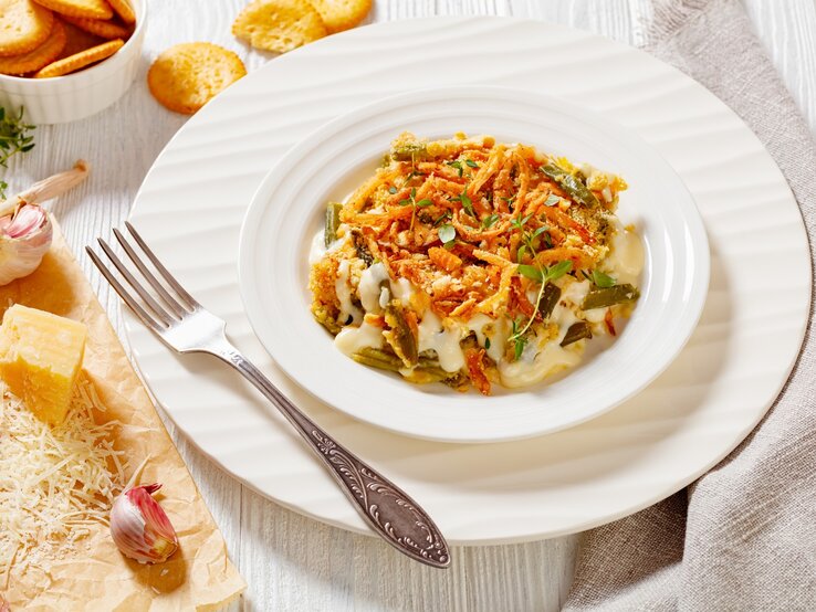 Bretonischer Bohnenauflauf auf weißem Teller mit Unterteller und Gabel auf hellem Untergrund. Im Bild Cracker, Parmesan und Knoblauch. Draufsicht.