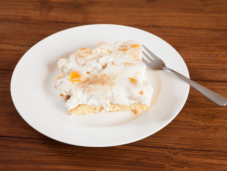 Ein angeschnittenes Stück Pfirsich-Schmand-Kuchen auf weißem Teller. 
