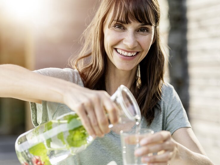 Frau mit braunen Haaren schüttet Wasser aus einer Karaffe in ein Glas | © Gettyimages/Westend61