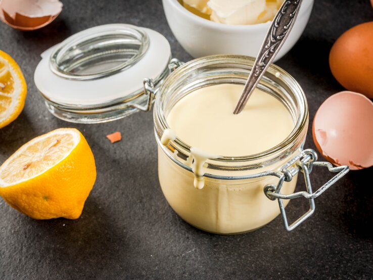 Ein Weckglas mit der Sauce hollandaise auf einem grauen Untergrund. Alles von schräg oben fotografiert und im Hintergrund sind noch die Zutaten erkennbar.