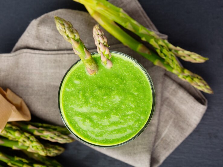 Grüner Smoothie mit Spargel in einem Glas. Darunter ein Küchentuch und daneben noch ein paar grüne Spargelstangen. 