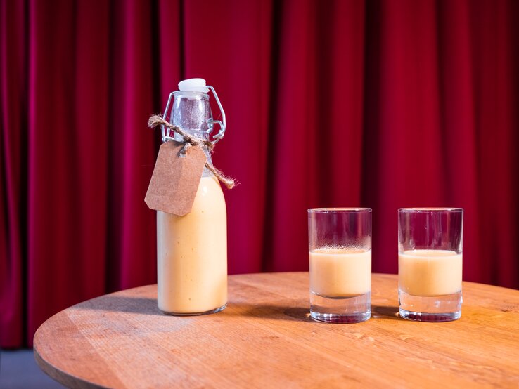 zwei Gläschen Eierlikör, daneben eine kleine Flasche. Das Ganze steht auf einem Holztisch, dahinter ein roter Vorhang.
