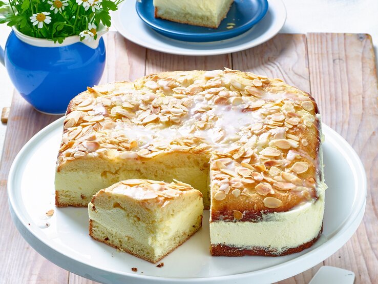 Der Bienenstich mit Zitronen-Mousse hat eine cremige Füllung und einen luftigen Teig. Die obere Schicht des Kuchens ist mit gerösteten Mandelsplittern und einem Zuckerguss bedeckt. Ein Stück des Kuchens wurde herausgeschnitten und liegt neben dem ganzen Kuchen auf der Tortenplatte. Im Hintergrund steht eine kleine Vase mit Blumen und ein Teller mit einem weiteren Stück Kuchen. Der Tisch ist rustikal aus Holz.