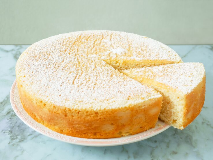 Runder, heller, angeschnittener Kuchen auf weißem Teller, bestäubt mit Puderzucker. 