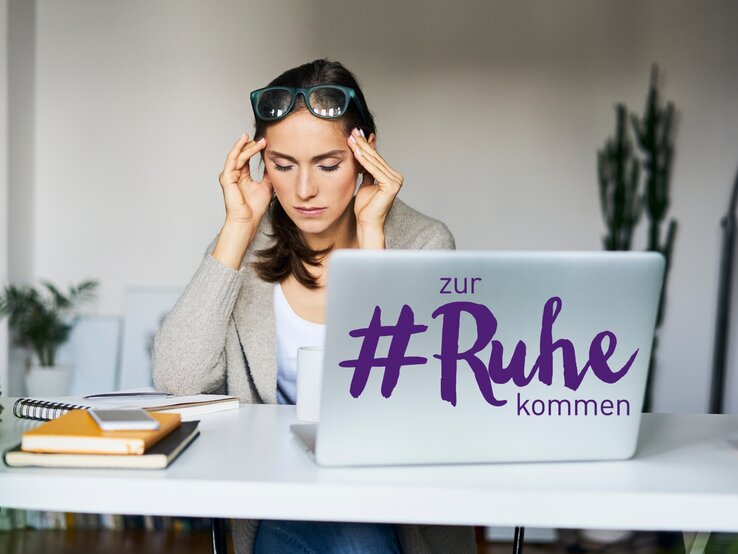 Eine junge Frau mit dunklen Haaren und einer Brille auf dem Kopf sitzt vor einem Laptop, auf dessen Deckel der Schriftzug "#zur Ruhe kommen" zu sehen ist. Sie hat die Augen geschlossen und ihre Finger an den Schläfen, was auf Stress oder Nachdenklichkeit hindeuten könnte. Vor ihr auf dem Tisch liegen ein Notizbuch, ein Tablet, ein Stapel bunter Haftnotizen und eine Tasse, was auf eine Arbeits- oder Lernsituation schließen lässt. Die Umgebung wirkt modern und hell.