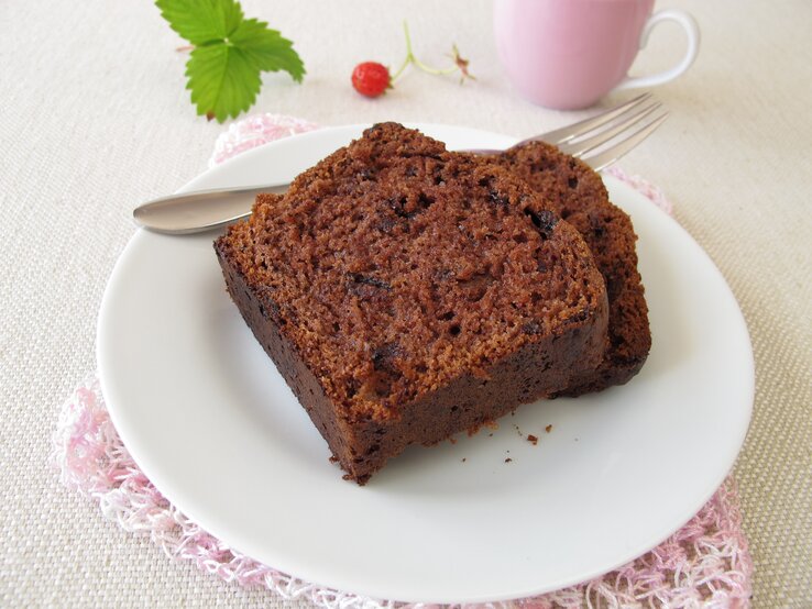 Zwei angeschnittene Stücke Schokoladenkuchen liegen auf einem weißen Teller. Daneben eine rosafarbene Tasse und ein Minzblatt. 