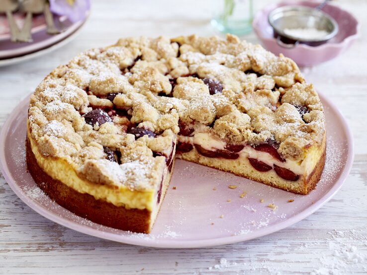 Ein rosa Teller mit einem Stück des Pflaumen-Puddingkuchens und Zimtstreuseln auf einem hellen Holztisch.