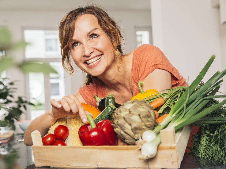 Eine strahlende Frau mit braunen, gelockten Haaren lehnt sich über eine Holzkiste voller frischer, bunter Gemüsesorten. Sie trägt ein korallenfarbenes Oberteil und zeigt mit einem Lächeln auf eine gelbe Paprika. Das Bild vermittelt eine fröhliche Stimmung und eine Leidenschaft für gesunde Ernährung. Sie befindet sich in einer modernen Küche, was auf eine häusliche und entspannte Atmosphäre hindeutet.