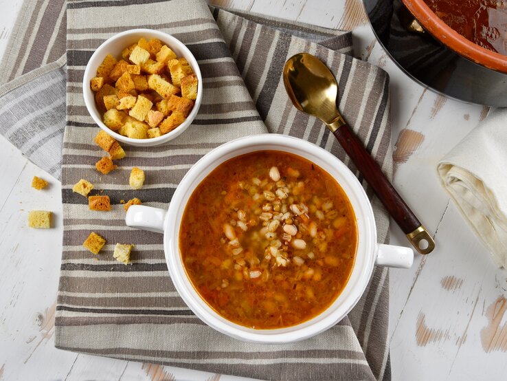 Draufsicht: Eine weiße Schüssel mit einer Kartoffelsuppe mit weißen Bohnen steht auf einem Küchenhandtuch, dass in verschiedenen Brauntönen gestreift ist. Daneben ist eine kleine Schüssel mit Brotwürfeln und einem Löffel. Drum herum stehen eine Schüssel dunkle Brühe sowie ein weißes Tuch.