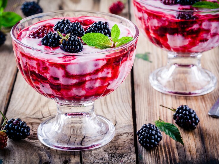 Süßes Löffelglück: eine unwiderstehliche Brombeer-Vanille-Creme Frontal: Untergrund aus Holz, zwei Dessert-Gläser Brombeer-Vanille-Creme und einige lose Brombeeren sowie ein Dessertlöffel.